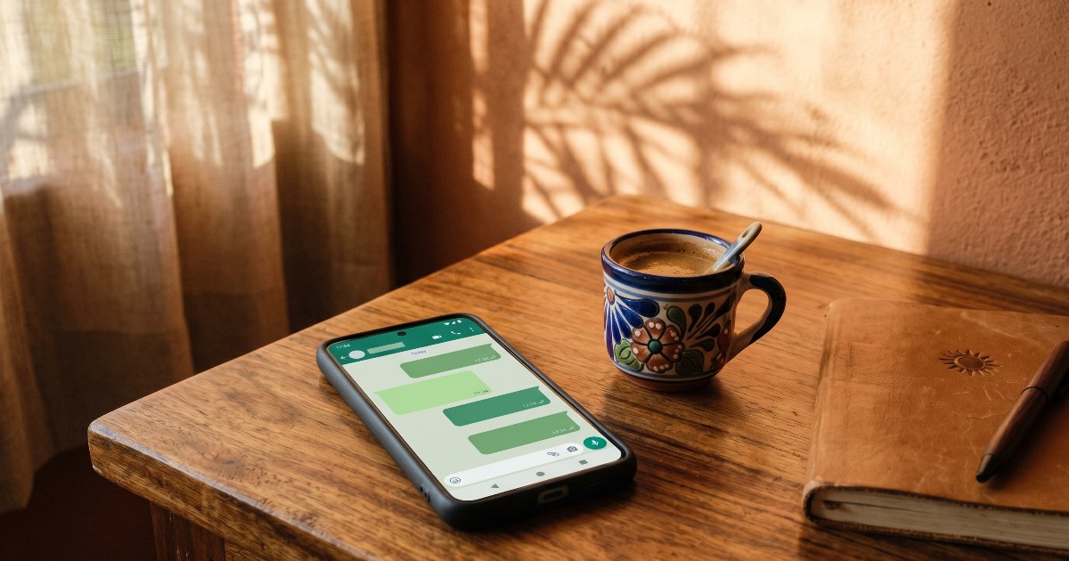 Smartphone sobre escritorio de madera con interfaz de chat, junto a taza de café mexicano y libreta de cuero, sombras de palmeras en la pared.